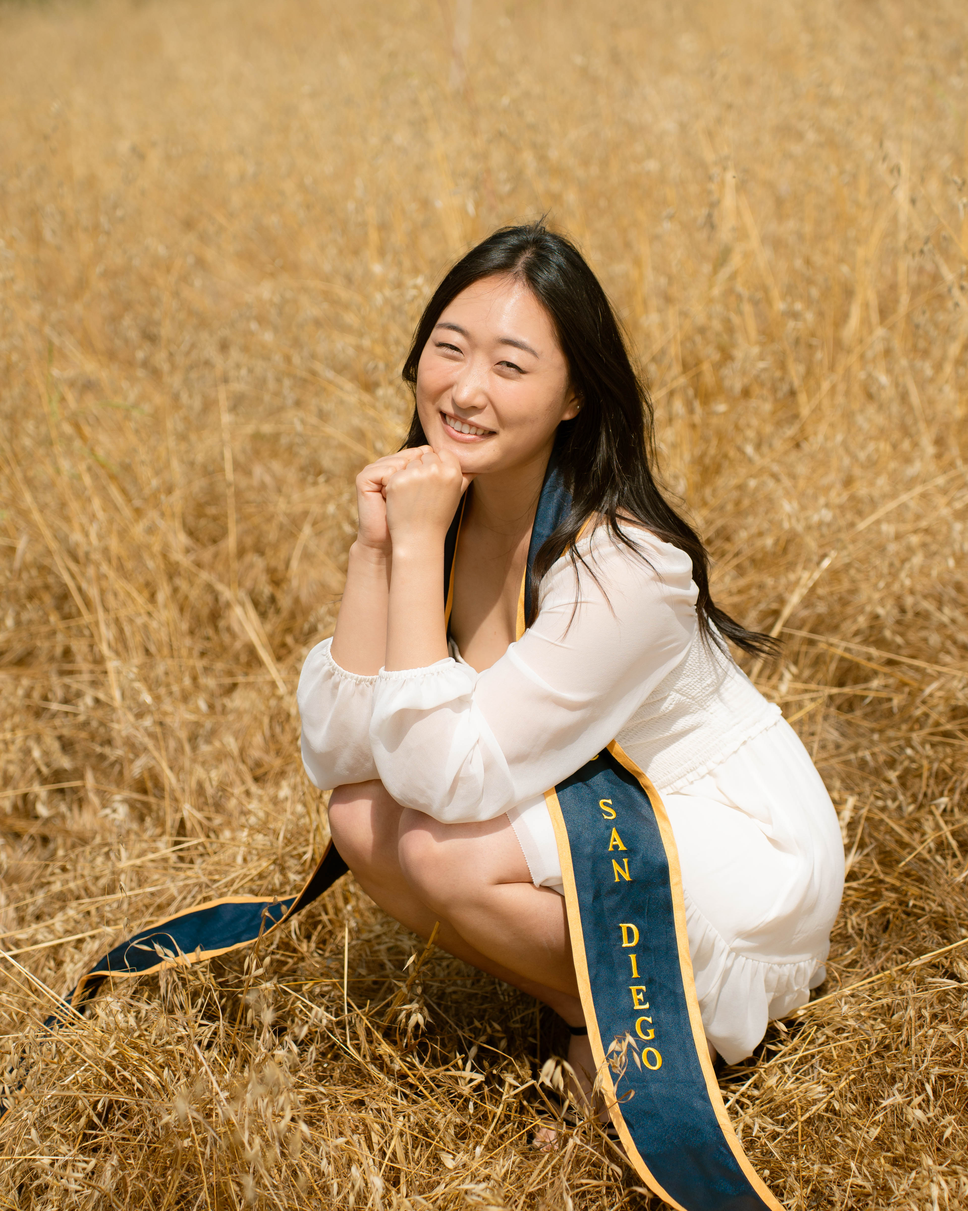 Kate sitting in a golden field wearing a white dress and a UC San Diego sash